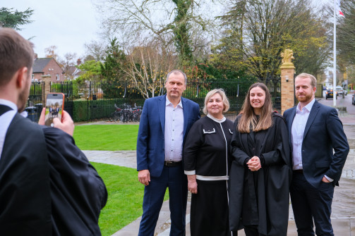 A group of four people pose outdoors for a photo on a cloudy day, with another person in academic attire taking their picture using a smartphone. The group stands on a path beside a green lawn, and one of them is wearing a graduation gown. Trees, bicycles, and a brick entrance gate are visible in the background.