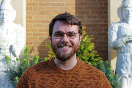 A man with brown hair, a beard and glass wearing a burnt orange jumper smiles outside the Lee Library