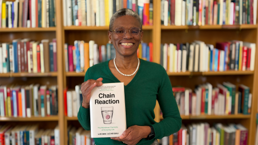 Smiling woman with short gray hair and glasses stands in front of library bookshelves while holding up the book “Chain Reaction: The Wondrous Chemistry of Everyday Life” by Ijeoma Uchegbu.