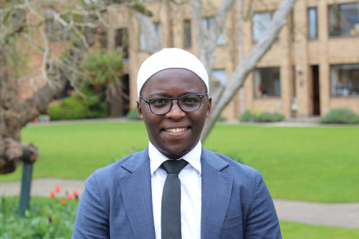 A smiling man wearing glasses, a white cap, a white shirt, black tie, and a blue suit jacket stands outdoors in front of a green lawn and brick buildings