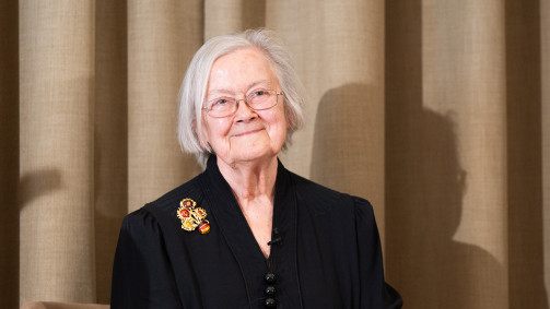 An older person with short white hair and glasses smiles gently while standing in front of beige curtains, wearing a dark formal outfit with a decorative brooch.