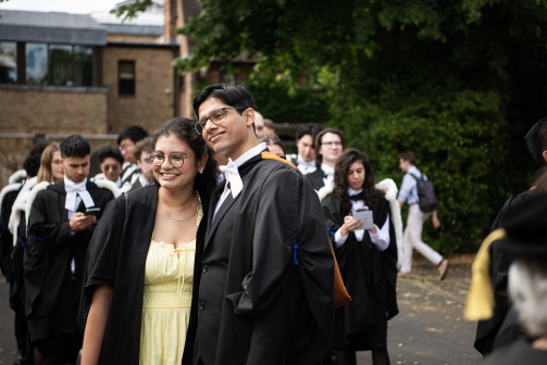 A smiling graduand in a black academic gown and white bow tie poses with a smiling companion in a light yellow dress. Other graduands in black gowns gather in the background, some looking at their phones, with trees and Wolfson buildings behind them