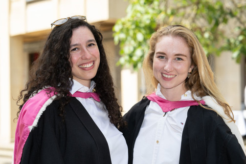 Two smiling graduates wearing black academic gowns with pink-and-white hoods stand side by side outdoors, posing for a portrait in front of a softly blurred building and green trees