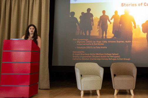 A woman stands at a red podium giving a presentation beside a large screen displaying a slide about refugee stories, with two empty chairs on the stage awaiting a panel discussion.