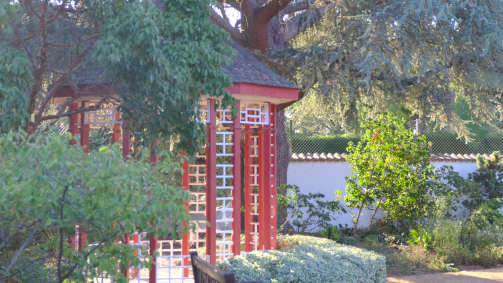 A red wooden garden pavilion stands among lush trees and shrubs, with sunlight filtering through the foliage in a peaceful landscaped setting.