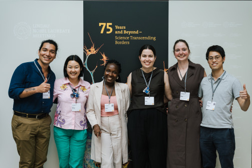 A group of six young scientists stand in front of a banner that reads '75 years and beyond - Science Transcending Borders', taken by Torben Nuding