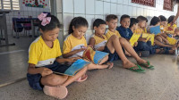 A group of young children in matching yellow and white shirts sit on the floor in a tiled room, reading books.
