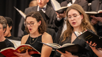 Two women in the foreground sing from open choral books while a larger choir stands behind them during a formal vocal performance.