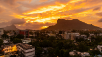 A cityscape at sunset with low-rise buildings and trees beneath a dark mountain silhouette, as vivid orange and gold clouds streak across the sky.