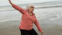 An older woman in a red striped shirt and glasses stretches her arms out while standing on a sandy beach with waves in the background.