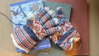 A pair of red, white, and blue patterned knitted gloves in progress with knitting needles and yarn rest on top of two books, one featuring a colorful bird and floral illustration on the cover.